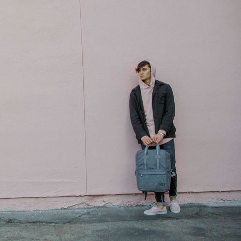 HEX Instinct Convertible Backpack in grey lifestyle view showing man holding backpack while leaning against a pink wall