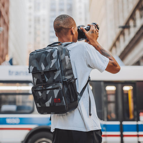 Man standing outside in front a bus wearing the Hex Glacier Camo Back Loader DSLR Backpack taking a picture